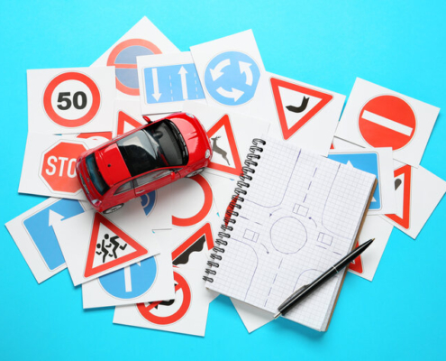 Viele verschiedene Verkehrszeichen-Karten, Notizbuch mit Skizze von Kreisverkehr und Spielzeugauto auf hellblauem Hintergrund