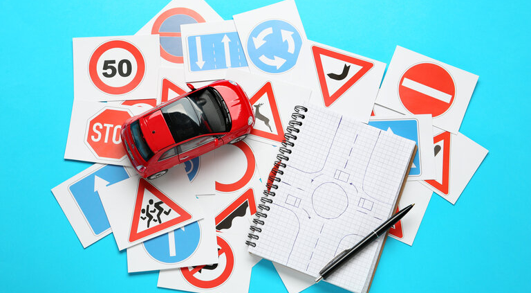 Viele verschiedene Verkehrszeichen-Karten, Notizbuch mit Skizze von Kreisverkehr und Spielzeugauto auf hellblauem Hintergrund