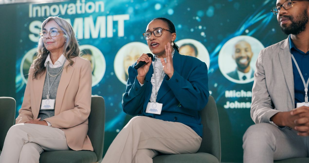 Eventmoderator bei einer Paneldiskussion auf einem Innovationsgipfel, Diskussionsteilnehmer mit Mikrofon vor modernem LED-Hintergrund