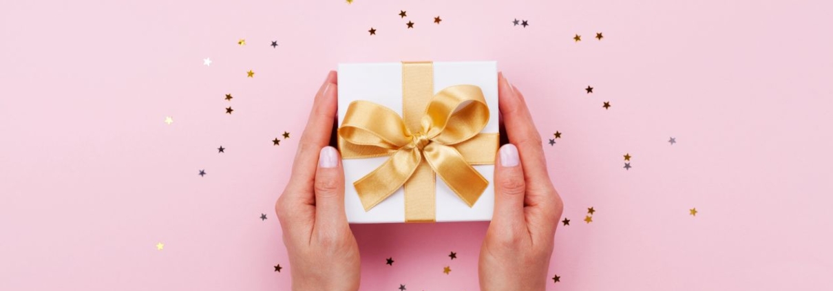 Zwei Haende halten eine elegante Geschenkbox mit goldener Schleife – stilvolle praesentkorb-Inspiration auf rosa Hintergrund