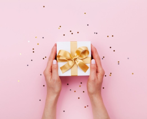 Zwei Haende halten eine elegante Geschenkbox mit goldener Schleife – stilvolle praesentkorb-Inspiration auf rosa Hintergrund