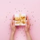 Zwei Haende halten eine elegante Geschenkbox mit goldener Schleife – stilvolle praesentkorb-Inspiration auf rosa Hintergrund