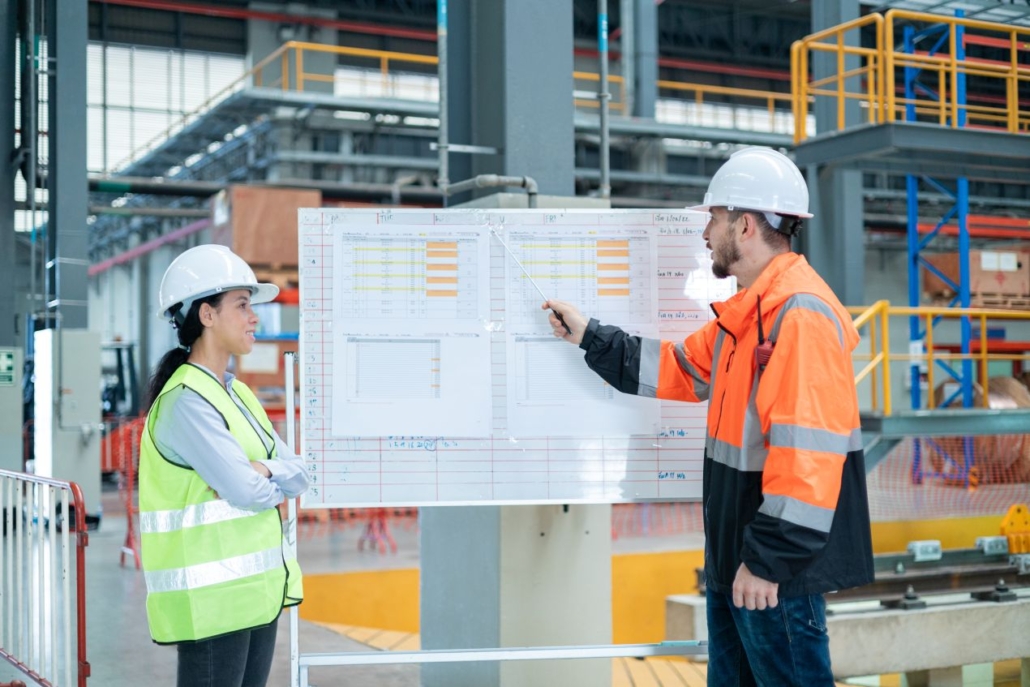 Zwei Mitarbeitende analysieren Prozessdaten an einem Whiteboard in der Produktionshalle – Beispiel fuer strukturierte Logistikplanung.
